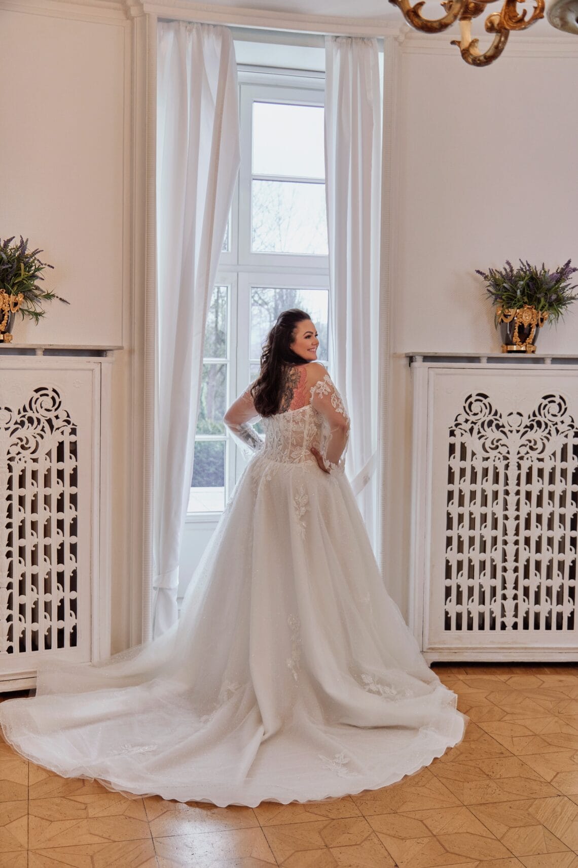 Braut in elegantes Hochzeitskleid vor Fenster in stilvollem Raum.