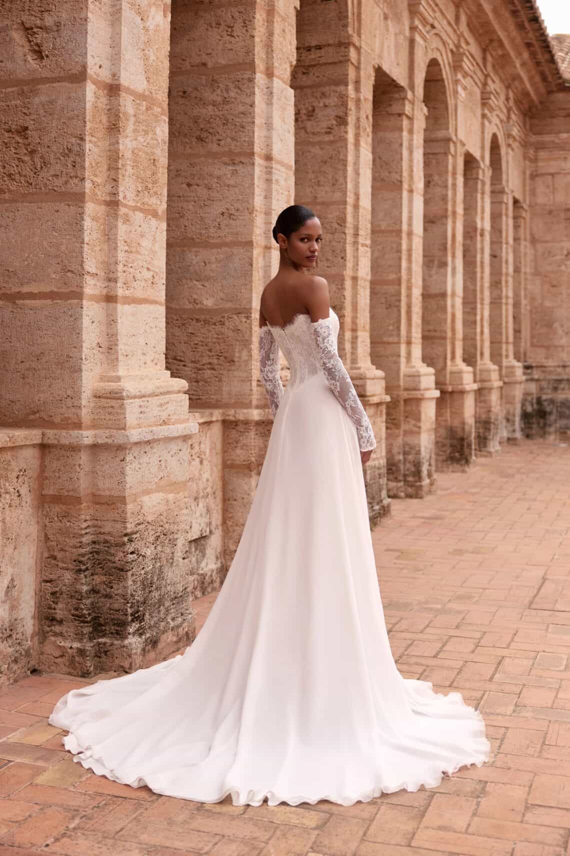 Elegantes Brautkleid mit Spitzenärmeln vor antiker Steinarchitektur.